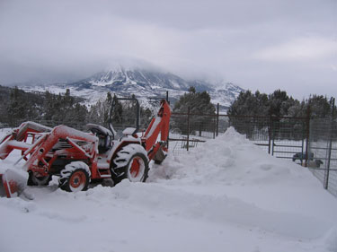 The tractor is parked outside in the snow, ready to plow the driveway. 