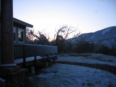 A light cover of snow covers the ground, the trees, and a nearby house as the sun rises. 