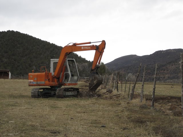 An excavator cleans brush and debris out from a drainage ditch running parallel to a fence.