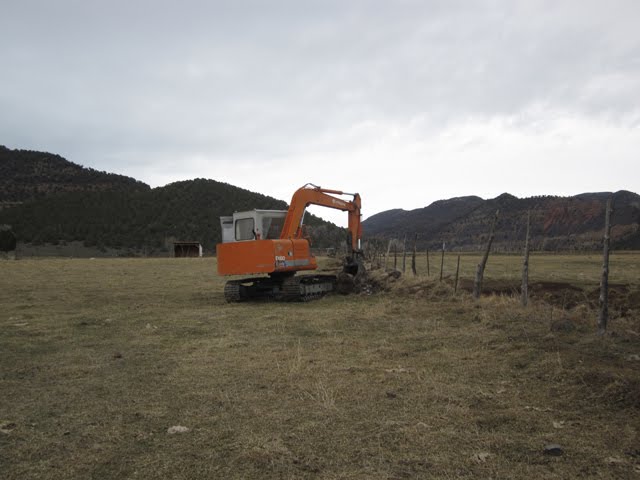 An excavator cleans brush and debris out from a drainage ditch running parallel to a fence.