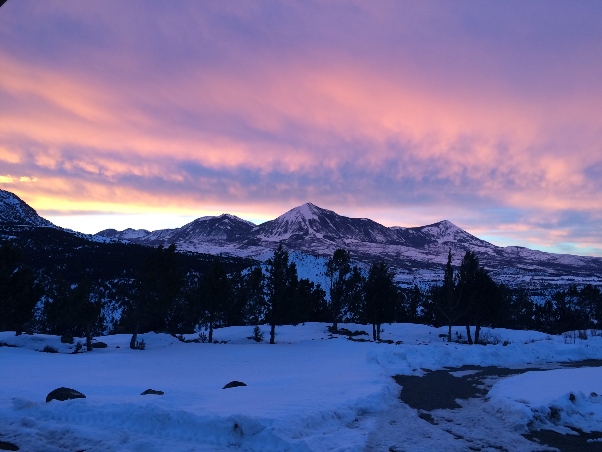 A pink and purple sunrise can be seen creeping over snowy mountains and trees. 