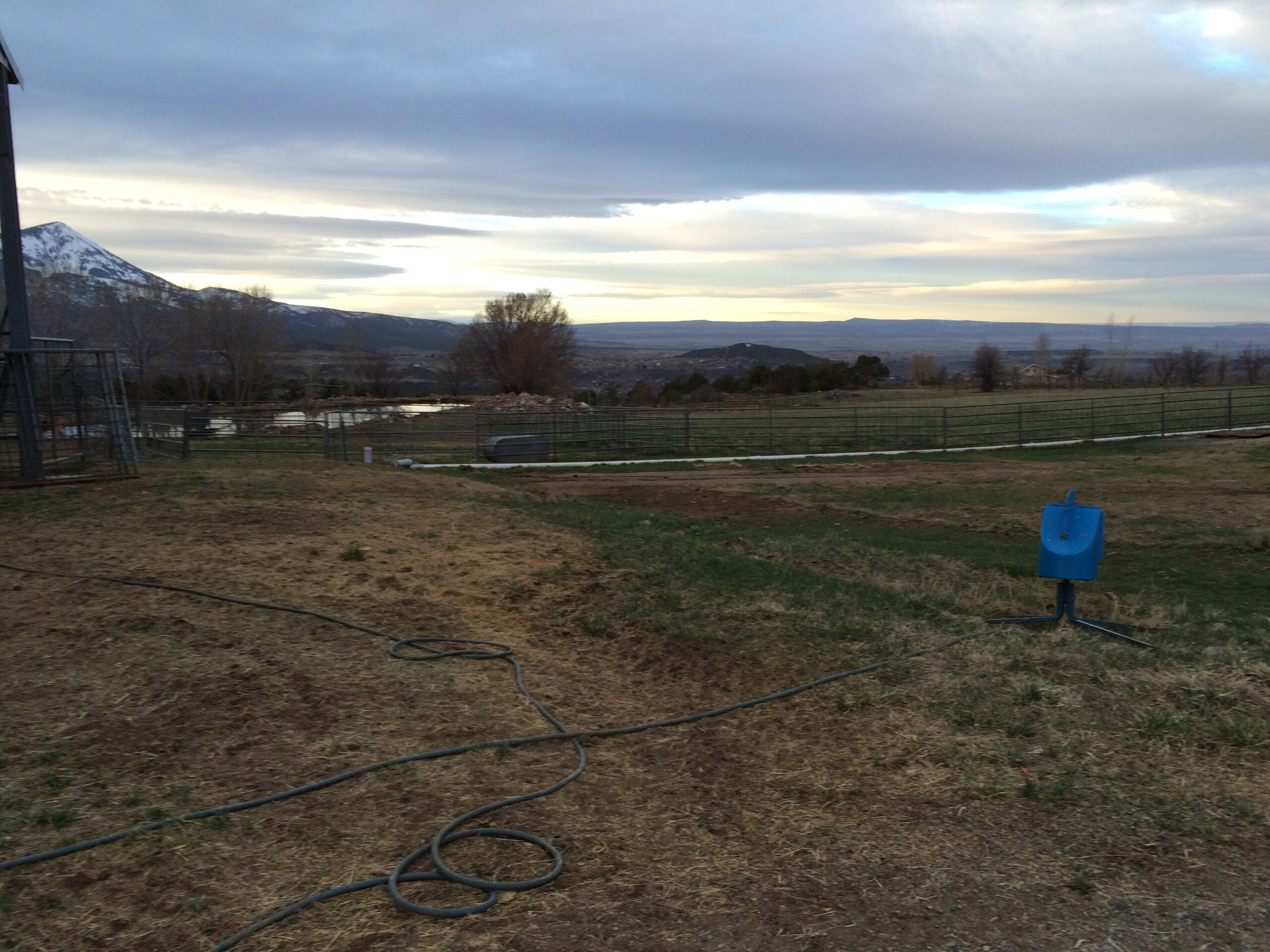 A view of an empty field without corrals, against a backdrop of mountains and trees. 