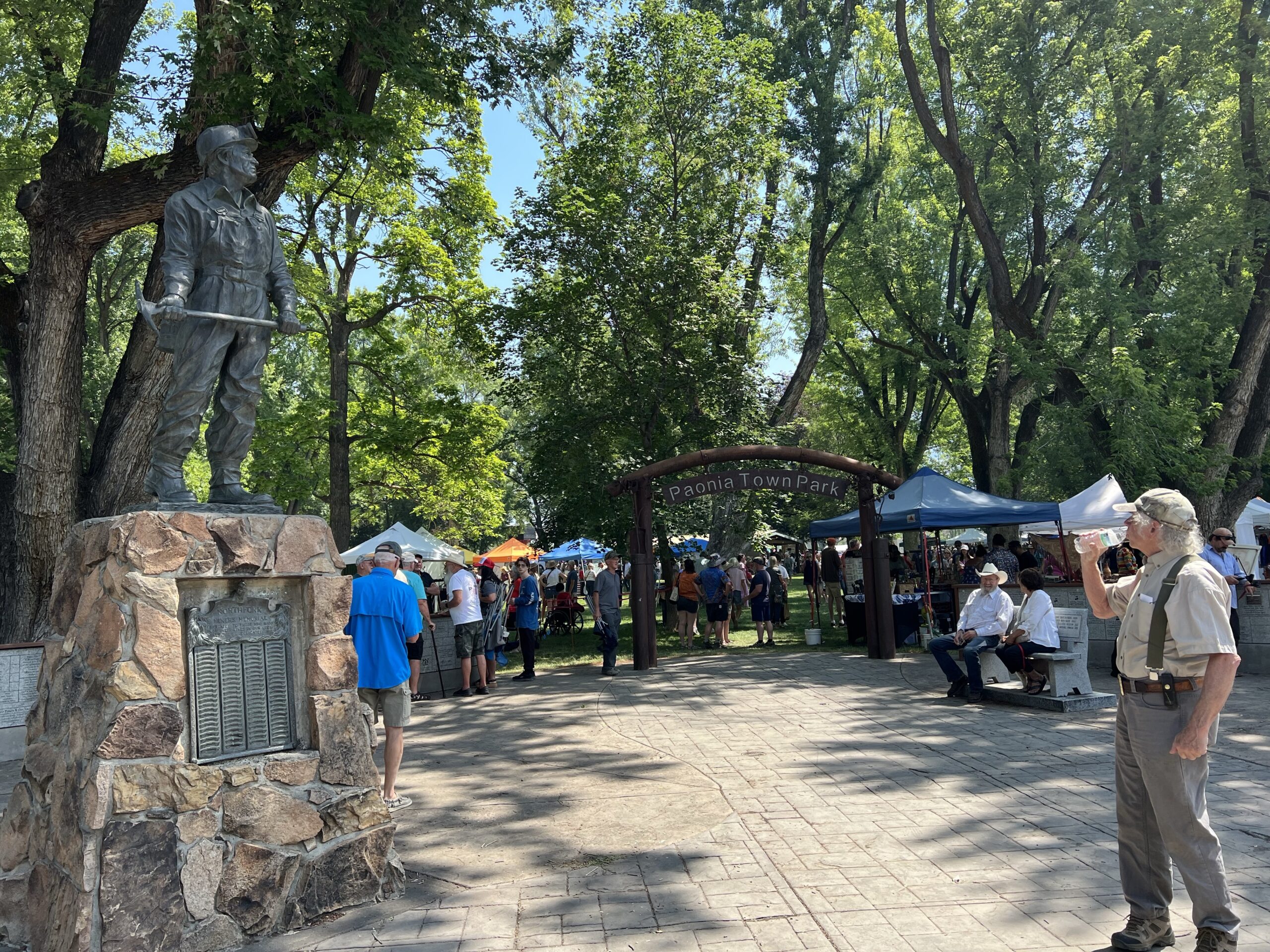 Many tents are set up in a green park full of trees, with a memorial statue visible in the foreground.