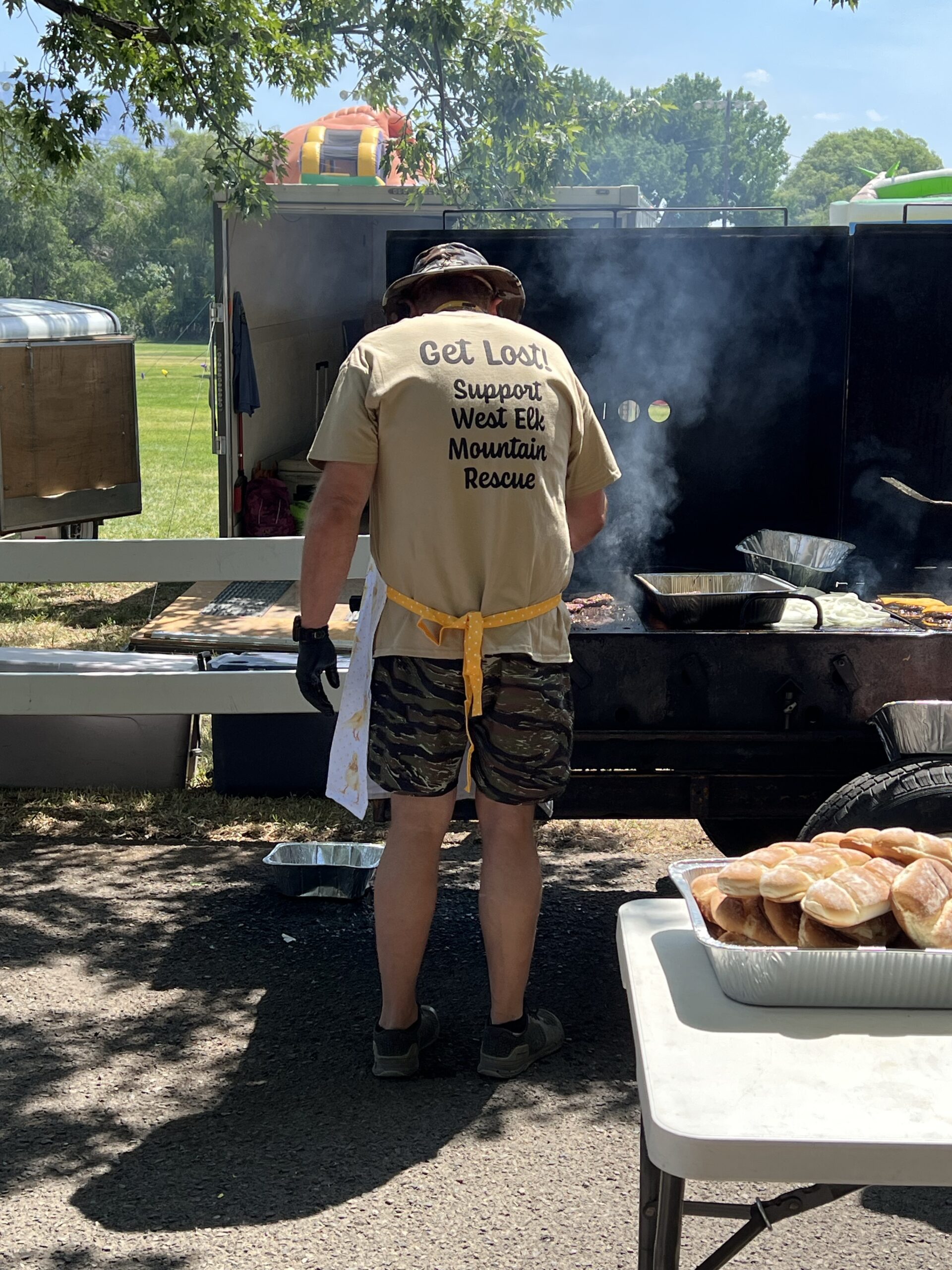 A man in a Support West Elk Mountain Rescue shirt grills hamburgers on a flat top.